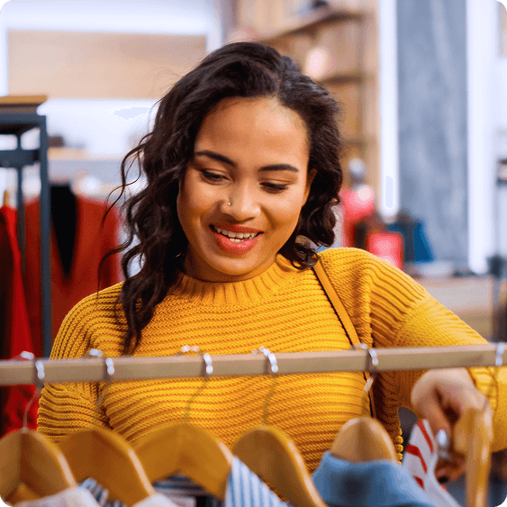 A smiling woman in a yellow sweater shops for clothes on a rack.