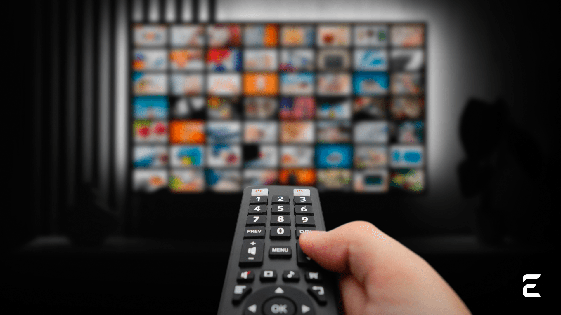 A hand holding a TV remote control in front of a blurred TV screen displaying a grid of content thumbnails.