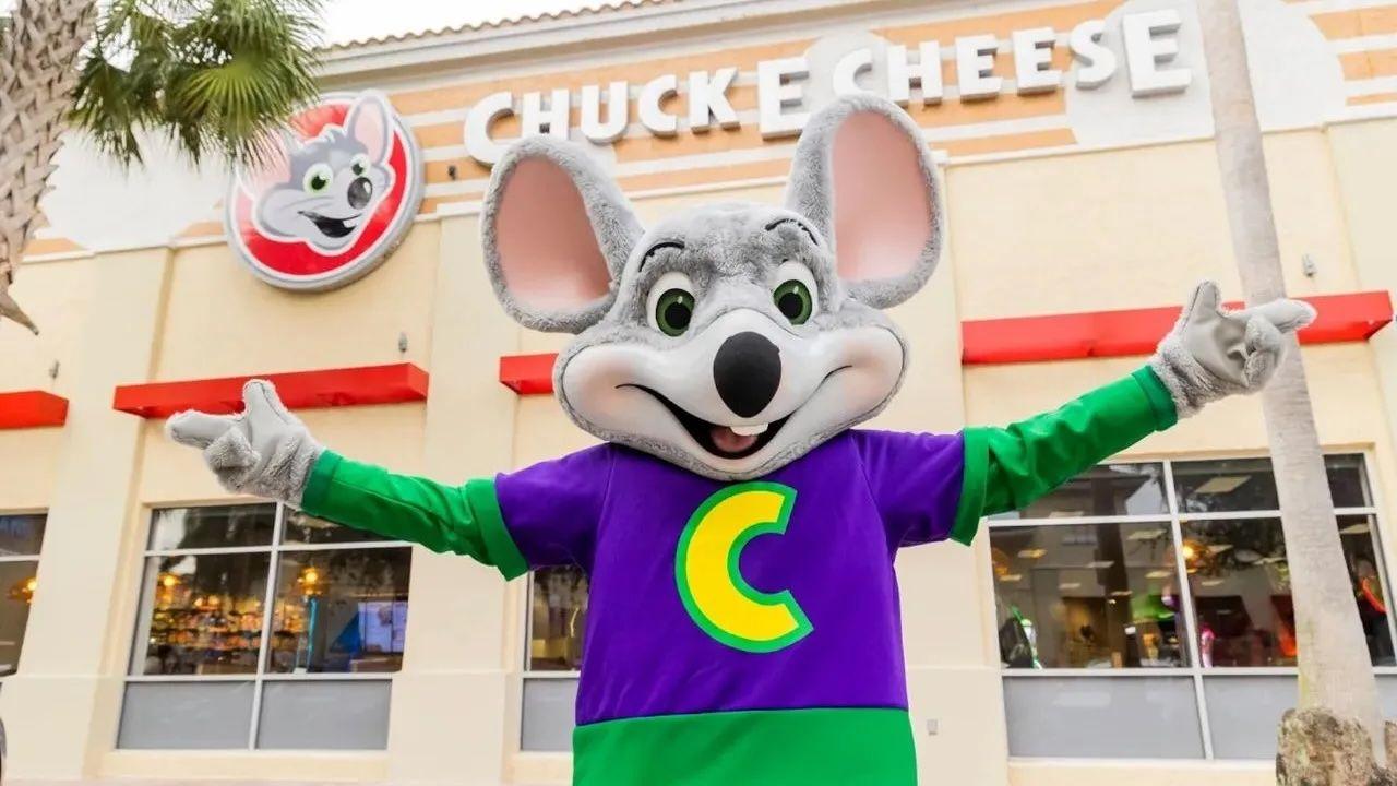 Chuck E. Cheese mascot in a purple shirt with a yellow "C", arms outstretched, stands in front of a Chuck E. Cheese restaurant.